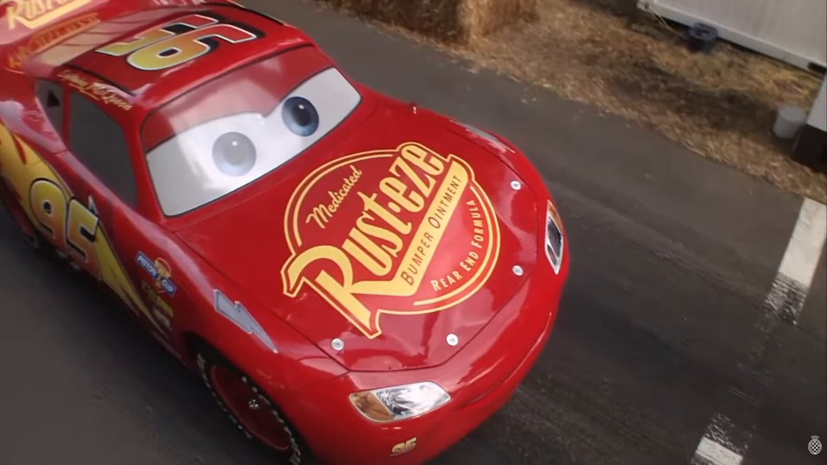 Vista superior del coche real animatrónico de Rayo McQueen rodando por el paddock del Goodwood Festival of Speed 2026, con balas de heno de fondo